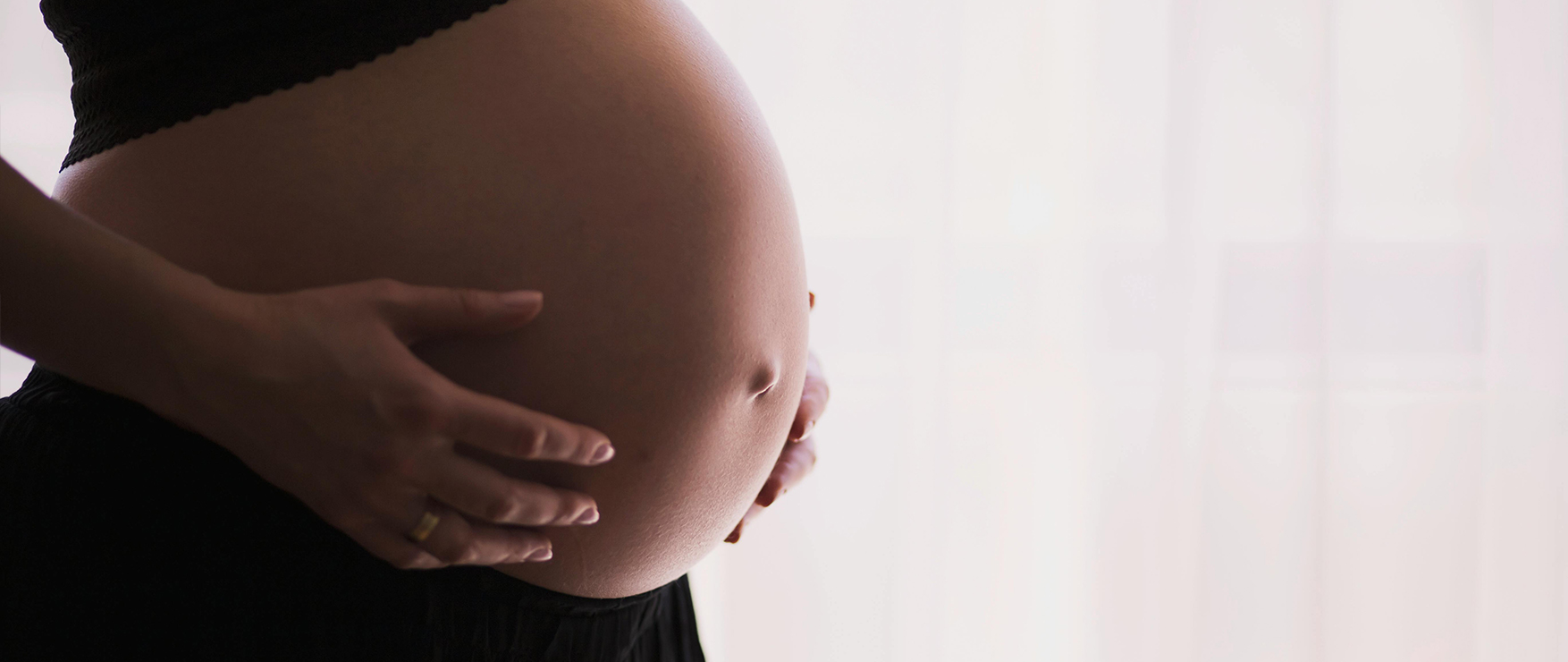 An image of a pregnant person's belly in front of a window with a sheer white curtain. This image is used as an introductory image for Solgar's blog, "8 Nutrients You Should Take During Pregnancy".