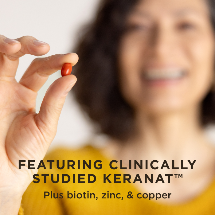 A woman smiling holding one red softgel of Solgar Hair Growth in her fingertips. Text on image reads, "Featuring clinically-studied Keranat, plus biotin, zinc, and copper"
