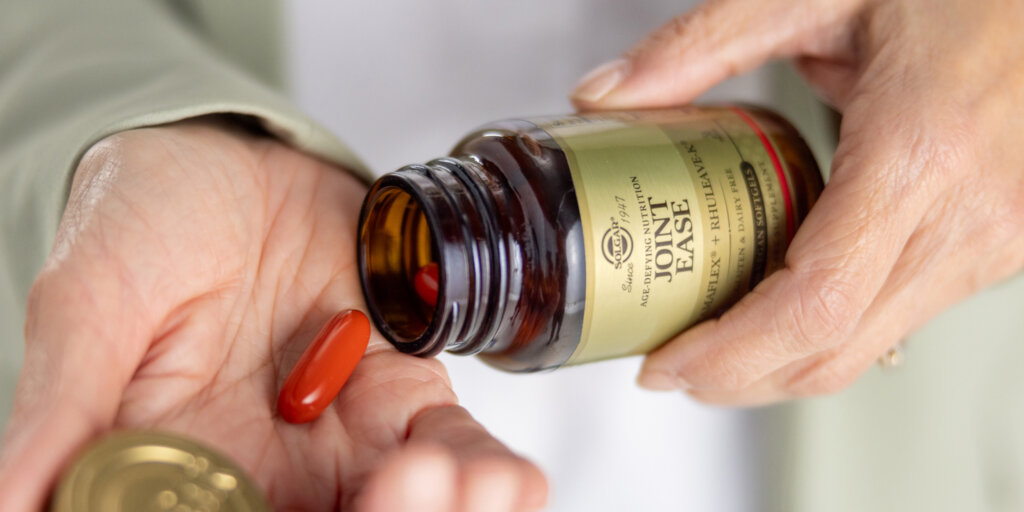 A closeup of an older woman's hands. She is holding a bottle of Solgar Joint Ease in one hand and pouring one capsule into her other hand. The capsule is red. 