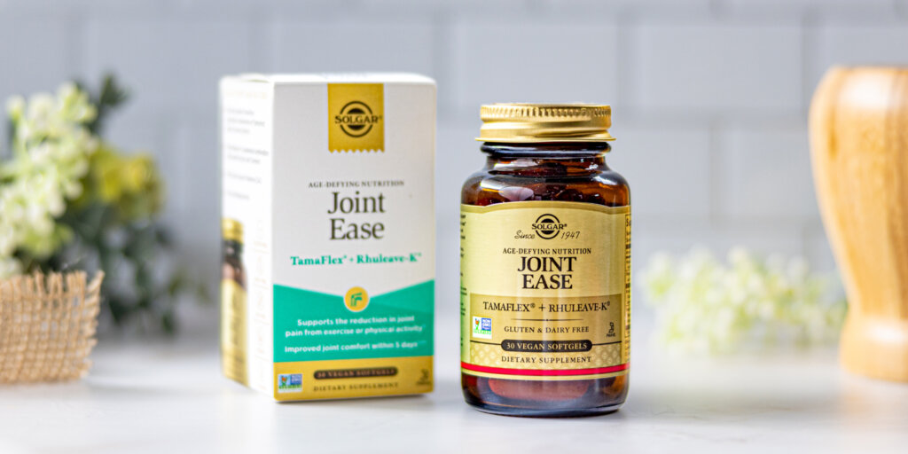 A box and amber glass bottle of Solgar Joint Ease on a clean, white kitchen scene. Some flowers and a wooden mortar and pestle surround the product. 