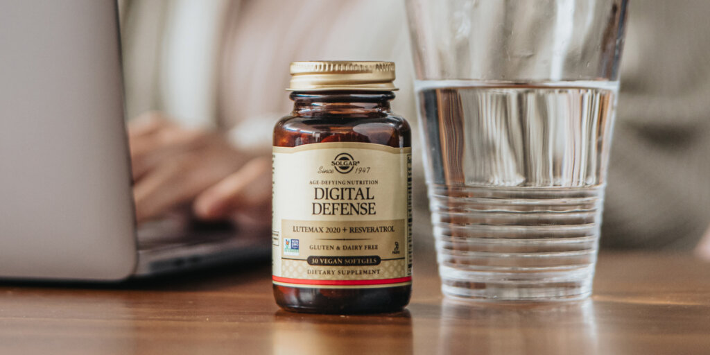 An amber glass bottle of Solgar Digital Defense capsules on a desk next to a glass of water. A woman is working at a laptop in the background. 