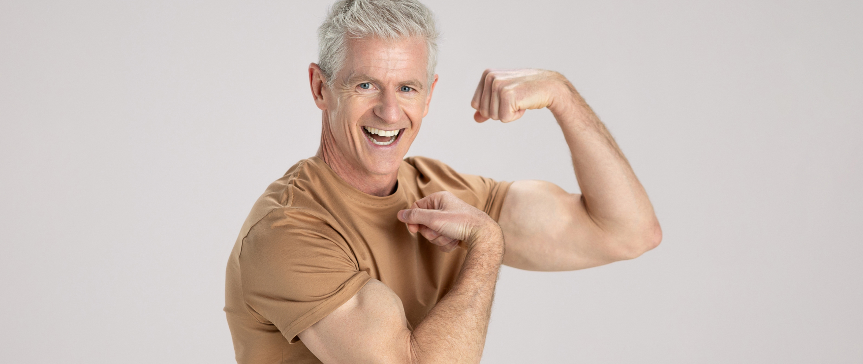 A fit, athletic man with grey hair flexes his bicep and is smiling at the camera.