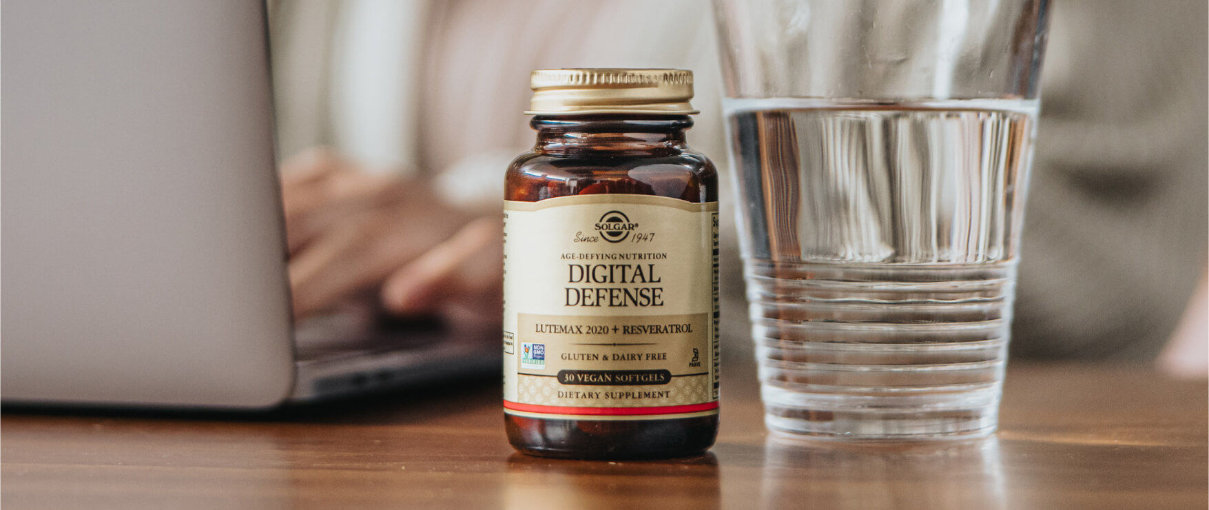 An amber glass bottle of Solgar Digital Defense Eye health softgels, next to a glass of water. A woman working on her laptop is in the scene.